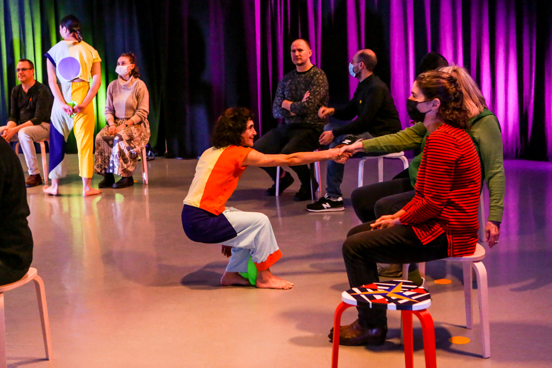 Choreographer Jody Oberfelder dressed in a color block costume of red, yellow, black, white and green, squats in front of a seated woman audience dressed in a long green shirt. They clasp forearms.